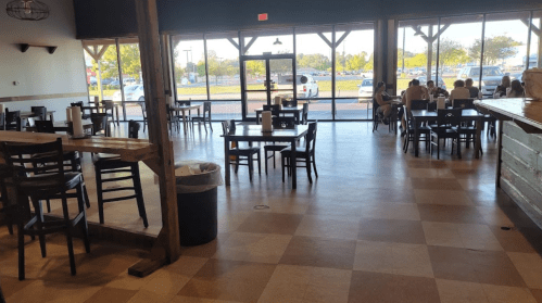 A spacious restaurant interior with tables and chairs, large windows, and a view of parked cars outside.