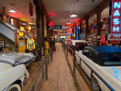 A vintage car museum featuring classic cars, neon signs, and retro decor along a wooden walkway.