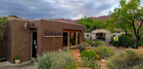 A rustic building with a sign reading "Sjava," surrounded by desert plants and mountains under a cloudy sky.