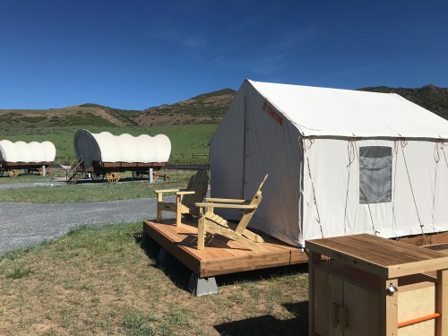 A white tent on a wooden deck with two chairs, surrounded by grassy fields and covered wagons in the background.