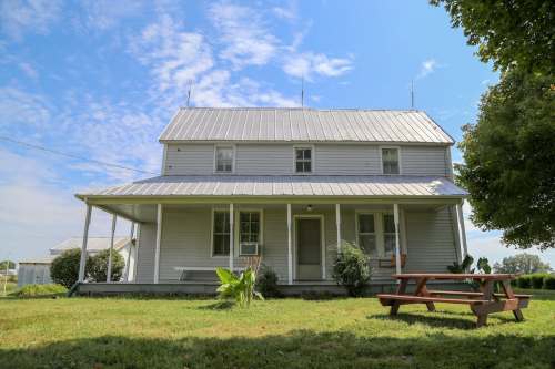 A two-story house with a metal roof, front porch, and picnic table on a grassy lawn under a blue sky.