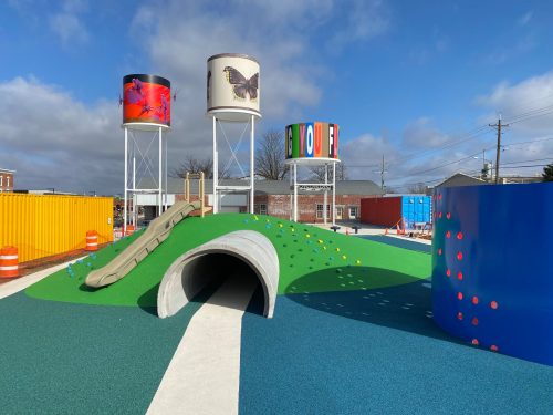 Colorful playground featuring a grassy hill, slide, and water tower art installations under a blue sky.