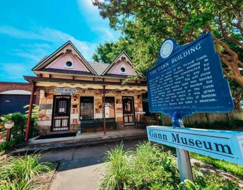 Historic Gann Museum building with a blue informational sign, surrounded by greenery and trees on a sunny day.