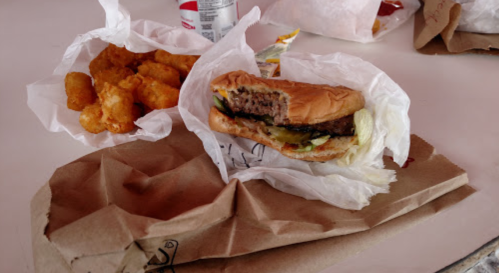 A close-up of a hamburger with lettuce and pickles, alongside a serving of golden tater tots on a table.