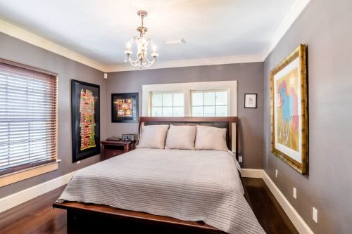 Cozy bedroom with a wooden bed, gray walls, a chandelier, and framed artwork. Natural light from windows.