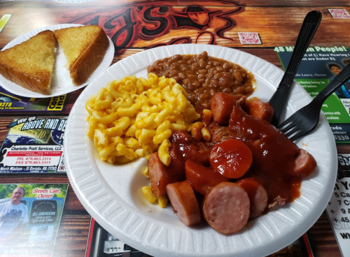 A plate with macaroni and cheese, baked beans, BBQ sausage, and toast on the side, served on a paper plate.