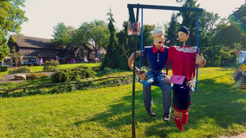 Two scarecrow figures sit on a swing in a sunny park, surrounded by greenery and a rustic building in the background.