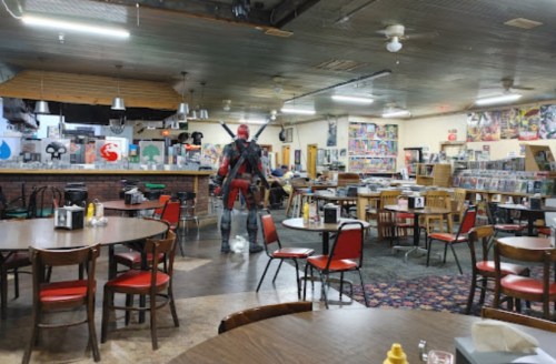 A person in a costume walks through a comic book-themed diner with tables and shelves filled with collectibles.