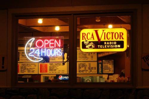 A brightly lit window displaying a neon "Open 24 Hours" sign and an RCA Victor advertisement, with a menu visible inside.