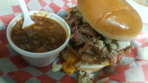 A pulled pork sandwich with cheese and coleslaw, served with a side of baked beans in a white cup.