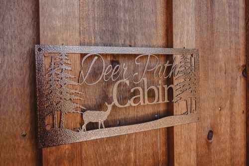 Metal sign reading "Deer Path Cabin" with trees and a deer design, mounted on a wooden wall.