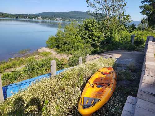 A bright yellow kayak rests on a path by a calm lake, surrounded by greenery and distant hills.