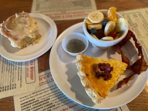 A plate with a slice of quiche topped with bacon, a bowl of fruit, a cinnamon roll, and a small cup of dressing.