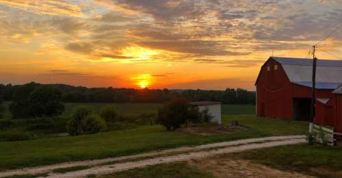A vibrant sunset over a rural landscape, with a red barn and a dirt path leading through green fields.
