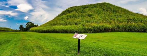 A grassy landscape featuring a large earth mound and an informational sign under a blue sky with clouds.