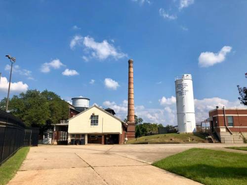 Industrial scene featuring a tall brick smokestack, water tower, and a building under a blue sky with scattered clouds.