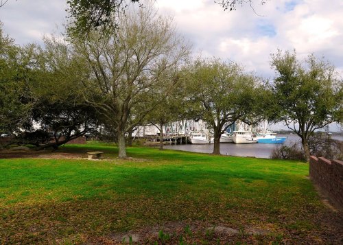A serene riverside scene with green grass, trees, and boats docked in the background under a cloudy sky.