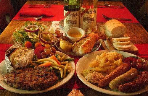A table set with two bottles of wine, plates of steak, lobster, sausages, vegetables, salad, and bread.