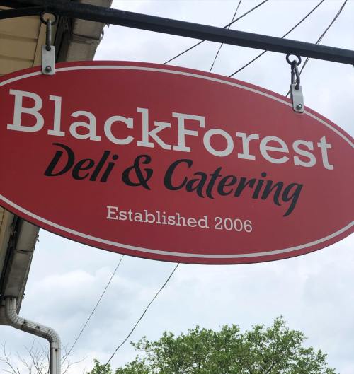 Sign for BlackForest Deli & Catering, established in 2006, hanging outside with a cloudy sky in the background.