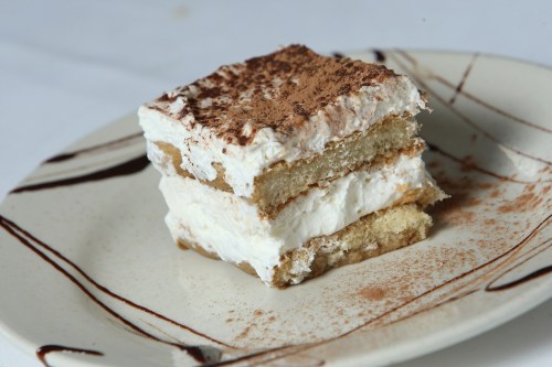 A slice of tiramisu on a plate, layered with cream and dusted with cocoa powder, drizzled with chocolate sauce.
