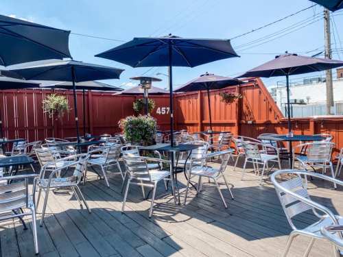 Outdoor dining area with metal tables and chairs, shaded by umbrellas, against a backdrop of a red fence.