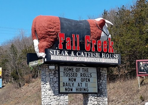 Sign for Fall Creek Steak & Catfish House featuring a large cow statue on top, advertising tossed rolls and hiring.