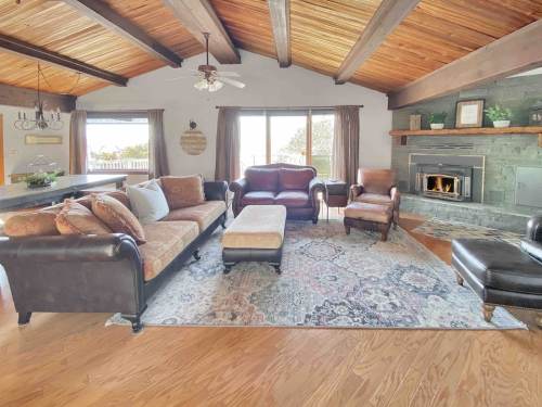 Cozy living room with wooden beams, a fireplace, plush seating, and a large area rug. Bright natural light from windows.