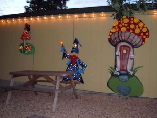 A whimsical outdoor scene featuring a painted wizard and mushroom on a yellow wall, with string lights and a picnic table.