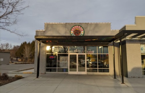 Exterior of a taqueria with a large sign, open door, and a cloudy sky in the background.