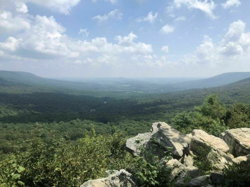 A panoramic view of lush green mountains and valleys under a partly cloudy sky. Rocks in the foreground.