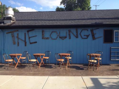 A blue wall with "THE LOUNGE" written in sticks, featuring several small tables and chairs outside.