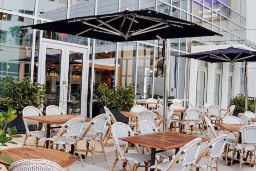 Outdoor dining area with wooden tables and chairs, shaded by large umbrellas, near a modern building.