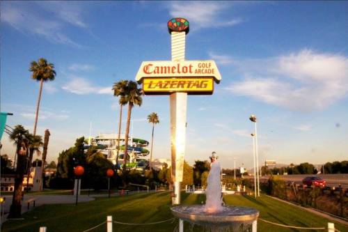 Sign for Camelot Golf Arcade and Lazer Tag, with palm trees and a fountain in the foreground under a blue sky.