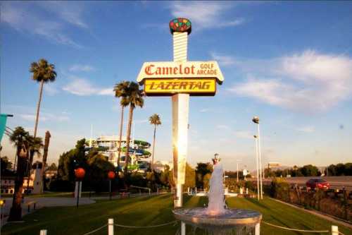 Sign for Camelot Golf Arcade and Lazer Tag, with palm trees and a fountain in the foreground against a blue sky.