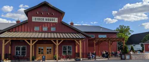 A red cider house with a wooden porch, surrounded by greenery and people enjoying the outdoor seating on a sunny day.