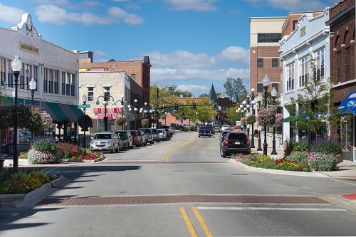 A charming downtown street lined with shops, parked cars, and colorful flower beds under a blue sky with fluffy clouds.