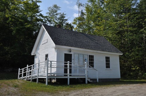 A small white house with a porch, surrounded by trees and greenery under a clear blue sky.