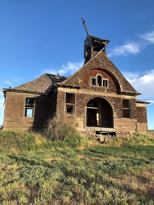 An abandoned, weathered wooden building with a steeple, surrounded by overgrown grass under a blue sky.