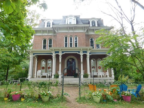 A large, historic brick house with a porch, surrounded by colorful flower pots and a lush garden.