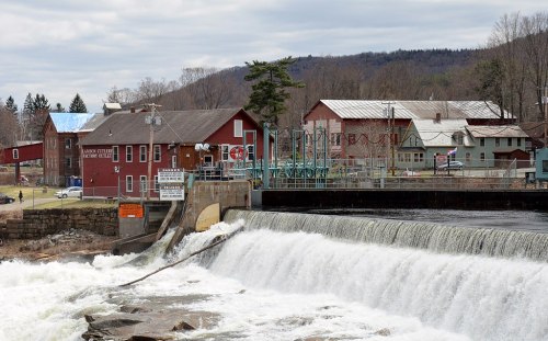 A scenic view of a dam with a waterfall, surrounded by buildings and trees under a cloudy sky.