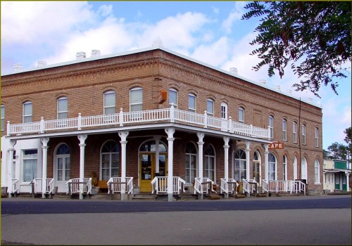 Historic brick building with a café sign, featuring a wraparound porch and white railings, set against a cloudy sky.