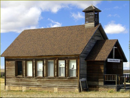 A rustic wooden building with a peaked roof and a small bell tower, set against a blue sky with clouds.