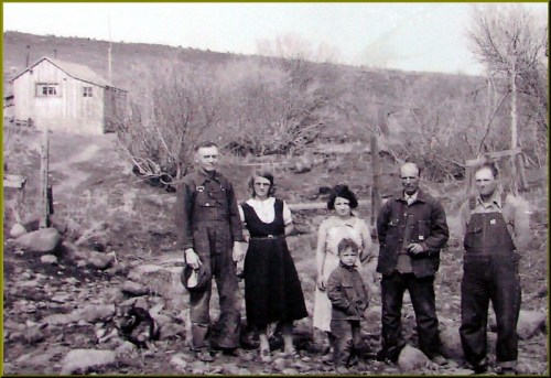A black-and-white photo of a family standing outside their rustic home, surrounded by trees and rocky terrain.