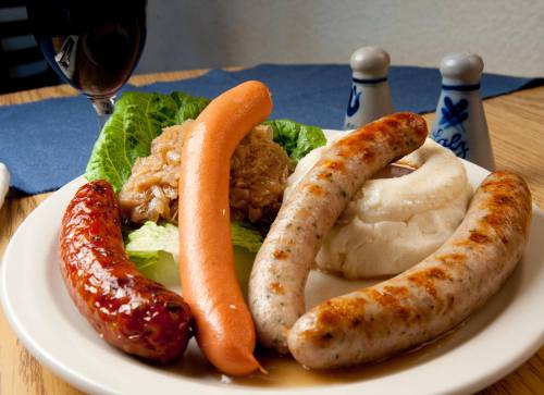 A plate of assorted sausages, mashed potatoes, lettuce, and sauerkraut, with a glass of red wine in the background.