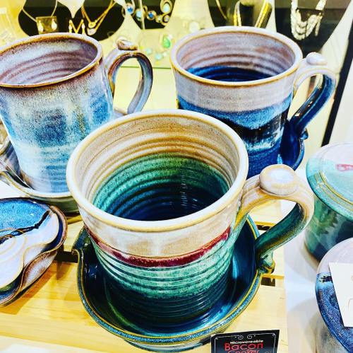 Three colorful ceramic mugs with unique glazes, displayed alongside jewelry in a shop.