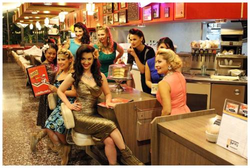 A group of women in vintage outfits poses at a retro diner, showcasing colorful dresses and hairstyles.