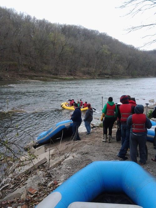 A group prepares for rafting on a river, with inflatable boats and lush green hills in the background.