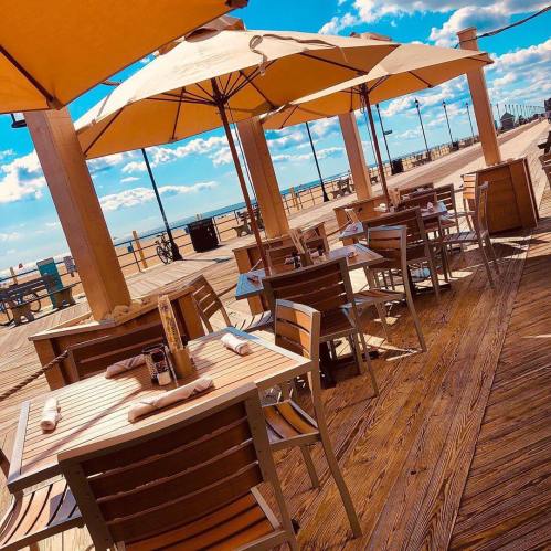 Outdoor dining area with wooden tables and umbrellas on a sunny boardwalk by the beach.
