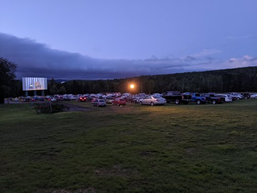 A drive-in movie theater at dusk, with cars parked in a grassy area and a large screen displaying a film.