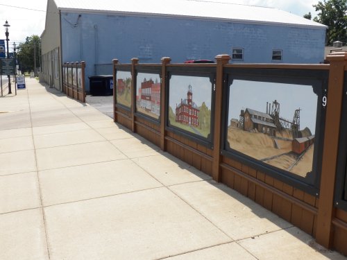 A sidewalk with a decorative fence featuring murals of historic buildings and industrial scenes.